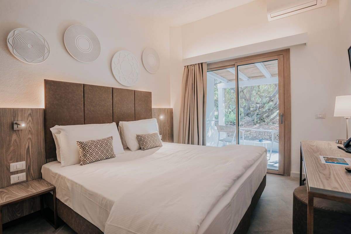 A modern hotel room with a large bed, a desk, and a balcony with a view of trees.