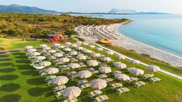 Un complexe hôtelier en bord de mer, avec des rangées de chaises longues et de parasols, un petit bâtiment et une vue sur la mer et les montagnes.