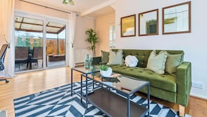 A modern living room with a green sofa, a glass coffee table, and a black and white rug. There is a sliding glass door leading to an outdoor area with a wooden deck.