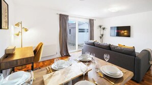 Een gemeubileerde woonkamer met een bank, een eettafel gedekt met borden en glazen, een aan de muur bevestigde televisie, en een schuifpui die leidt naar een terras.
