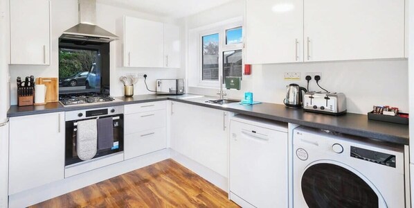 A modern kitchen with white cabinets, a stove, a dishwasher, and a washing machine. - Cozy Family Home | Pet-Friendly, Parking & Netflix (Farnborough)