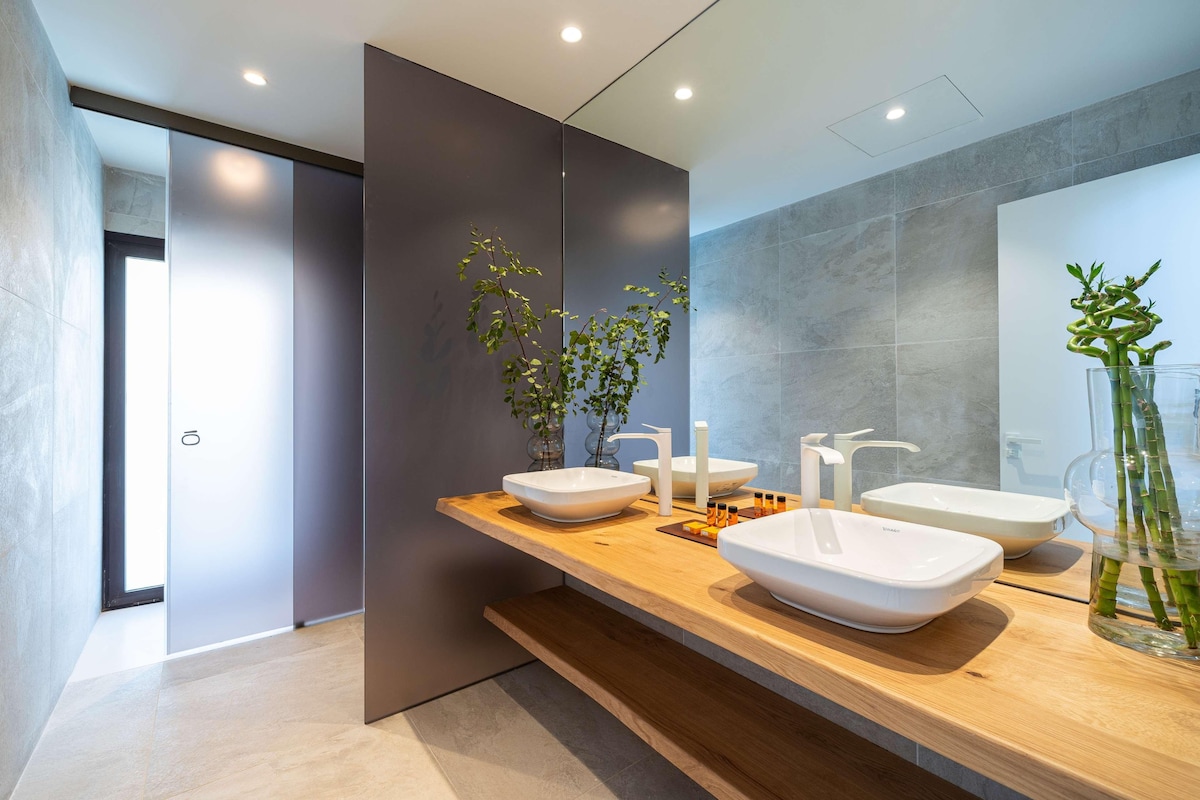 A modern bathroom with a wooden vanity, three white vessel sinks, a mirror, and a potted plant.