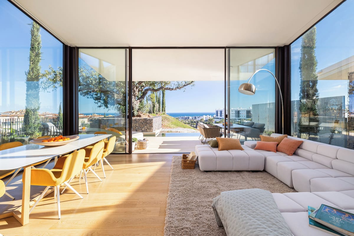 Una sala de estar moderna con un amplio ventanal, un sofá blanco, una mesa de comedor con sillas y vistas a la ciudad y al mar.