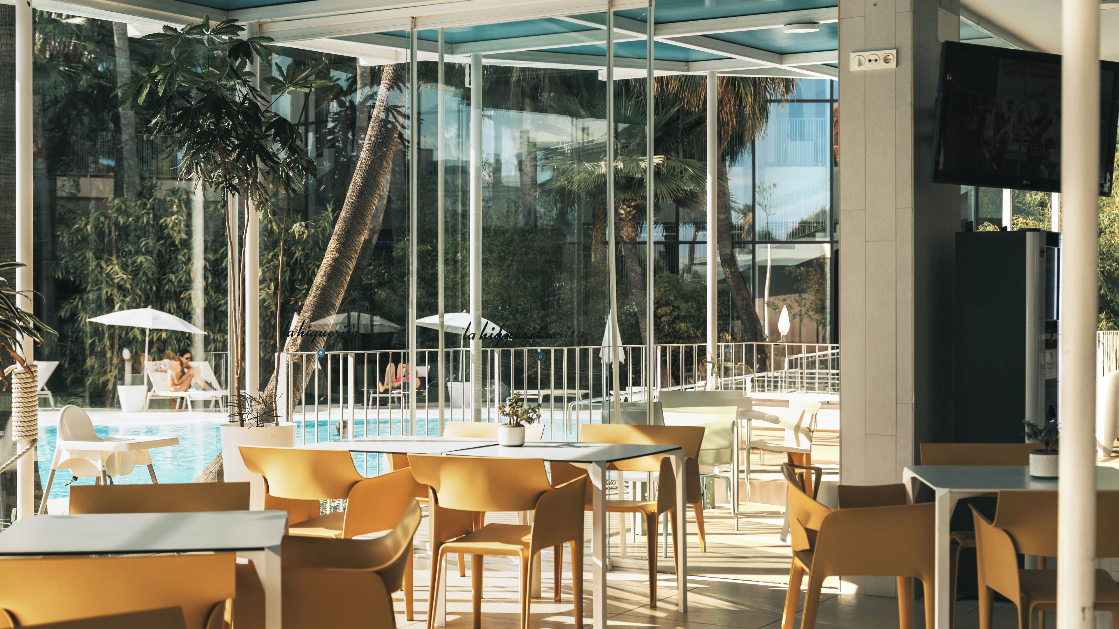 Un espace aménagé au bord de la piscine, avec des tables et des chaises, une véranda en verre et une vue sur la piscine et les palmiers.