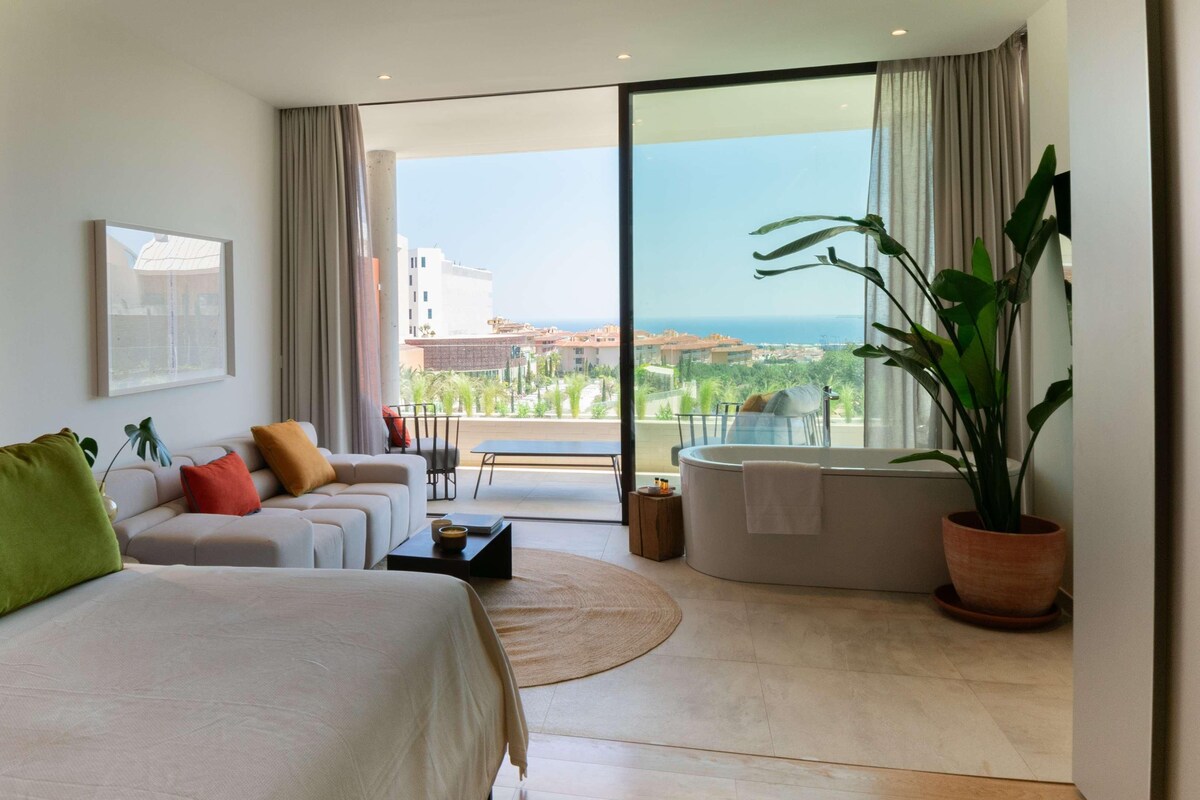 A modern bedroom with a large bed, a sofa, a small table, a bathtub, and a balcony with a view of the sea.