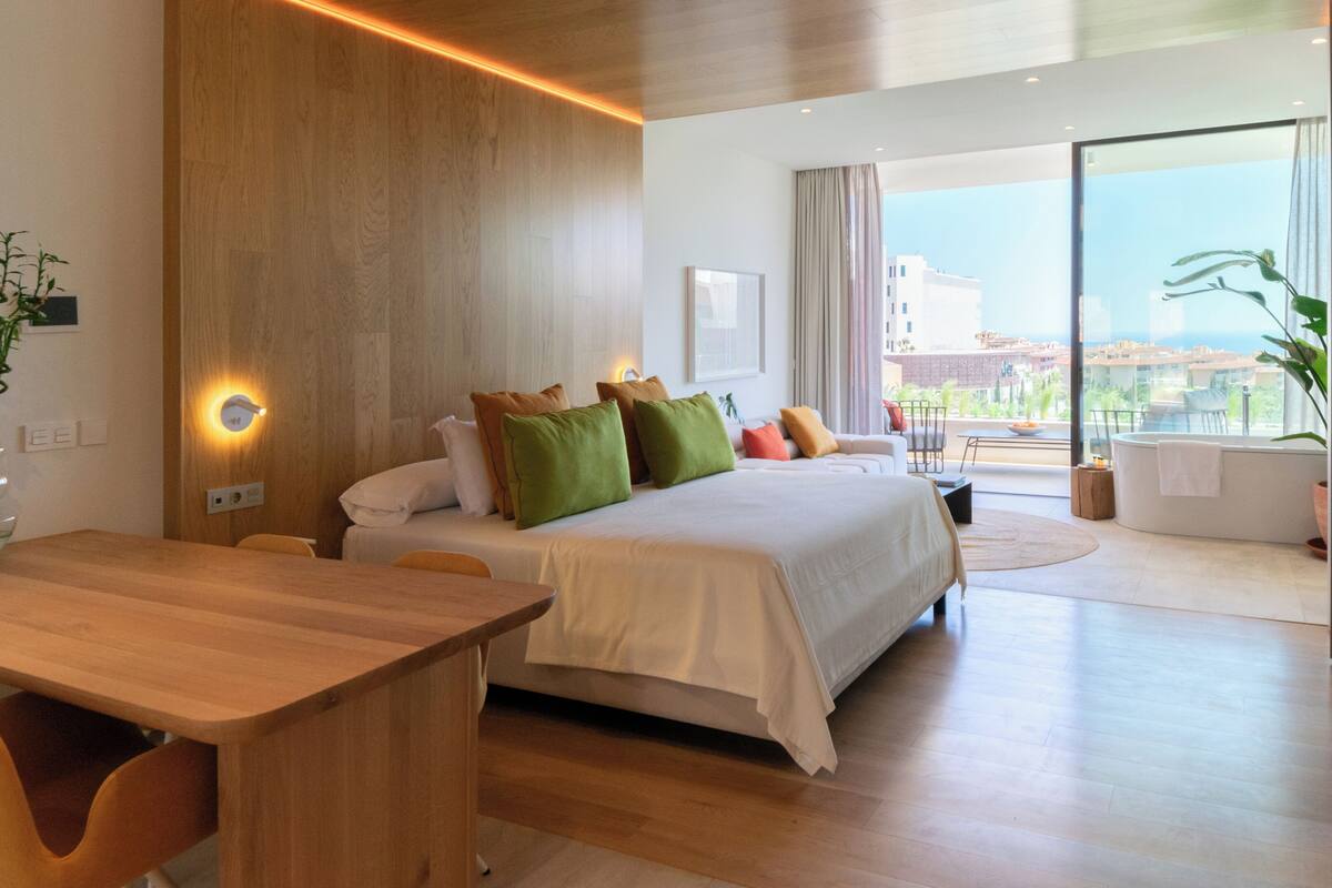 Una habitación de hotel moderna con una pared de madera decorativa, una cama grande con muchas almohadas, un escritorio de madera y vistas a la ciudad a través de amplias ventanas.