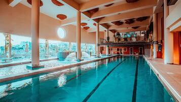 An indoor swimming pool with clear water, surrounded by white columns and a view of the city.