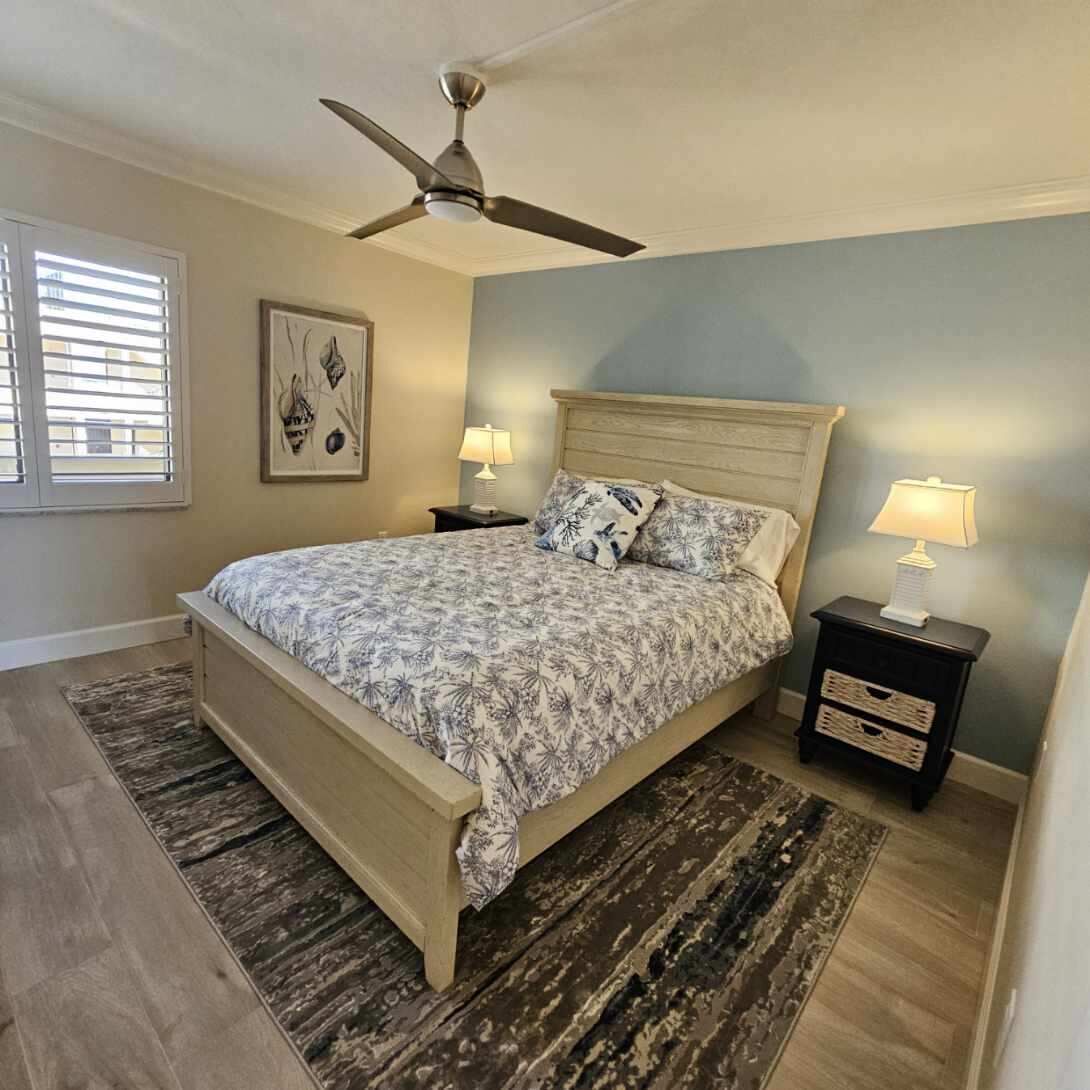 A bedroom with a bed, a ceiling fan, two bedside tables with lamps, a window with blinds, and a wall with a framed picture.