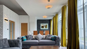 A modern living room with a grey sectional sofa, wooden coffee table, and yellow curtains. - Riverside Retreat - One Bedroom Cottage, Sleeps 2 (Deganwy)