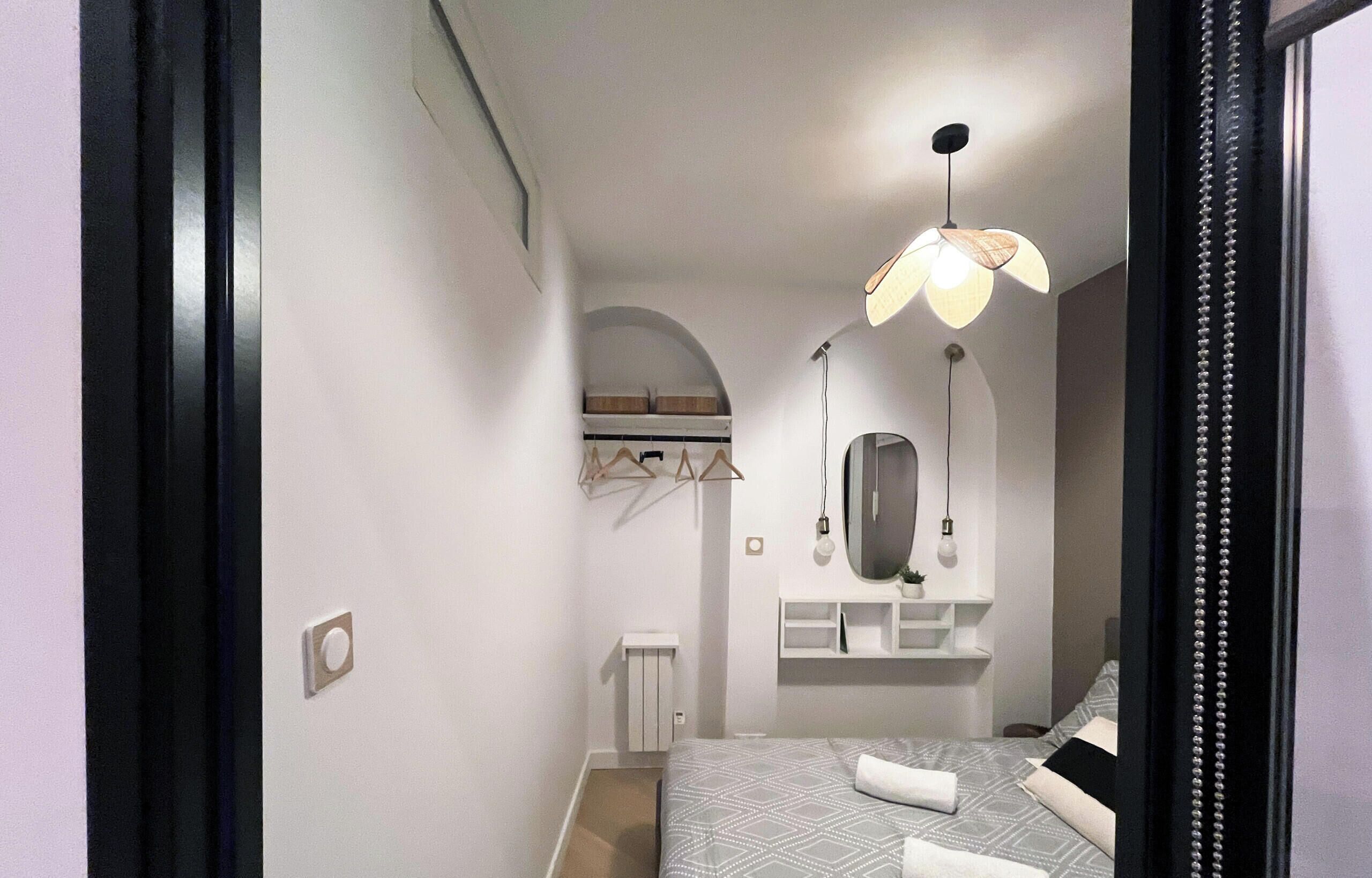 Une chambre à coucher moderne avec un lit, un miroir, un luminaire suspendu et une petite étagère sur laquelle est posée une plante.