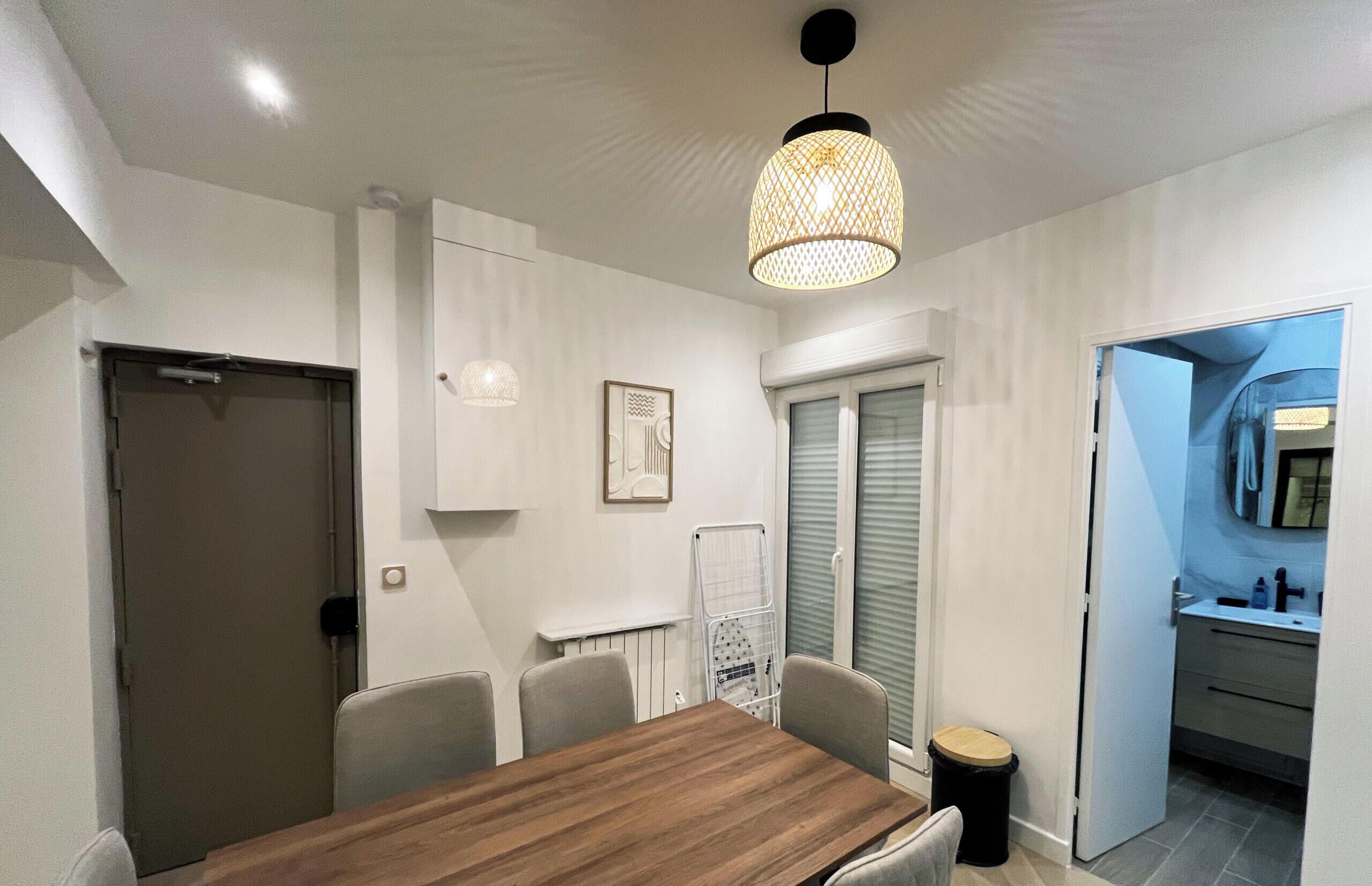 A modern dining area with a wooden table and chairs, a hanging light fixture, and a view into a kitchen area.