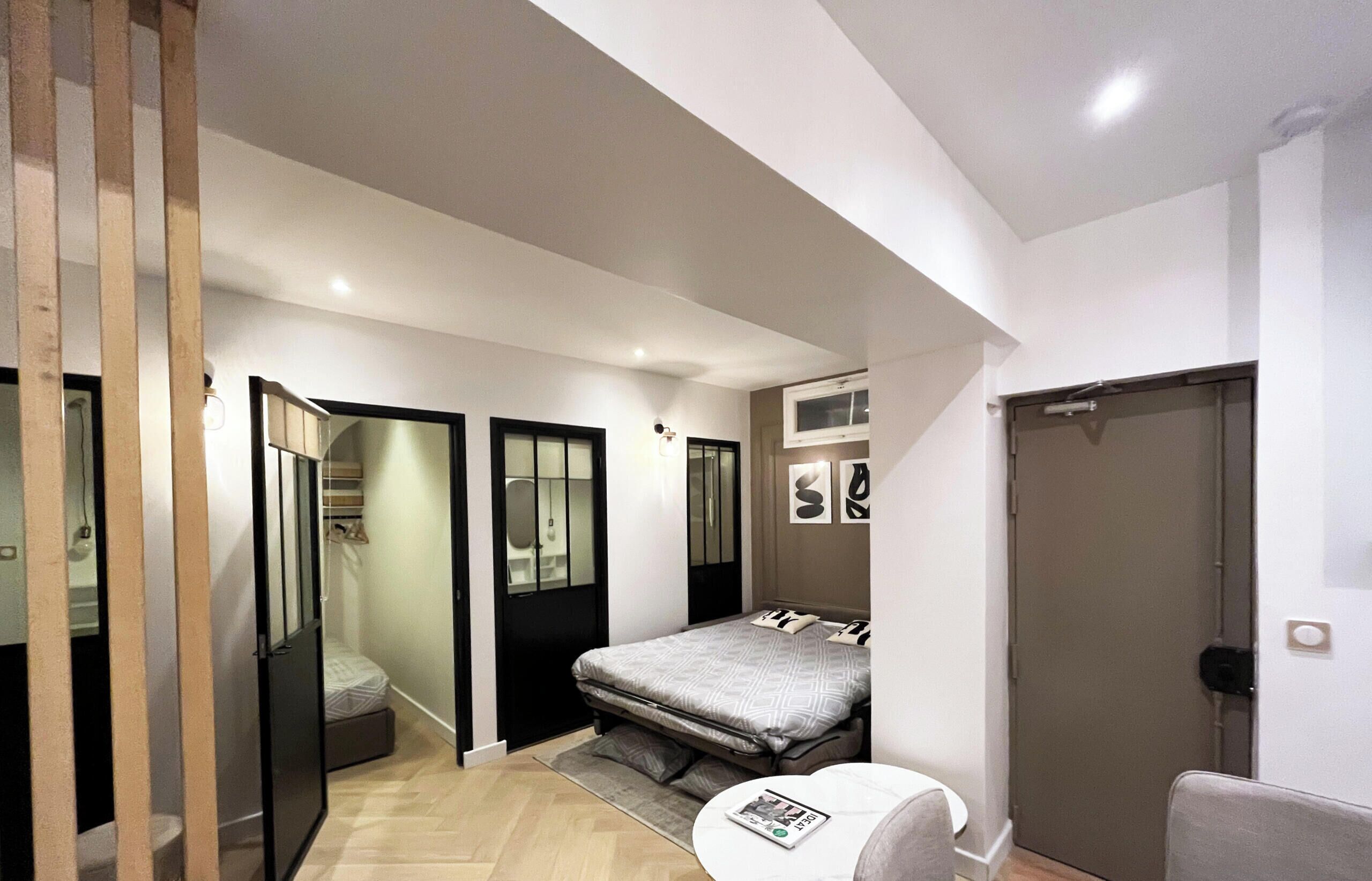 A compact bedroom with a bed, a small table, and a chair. There is a wardrobe with a mirror and a door leading to another room.