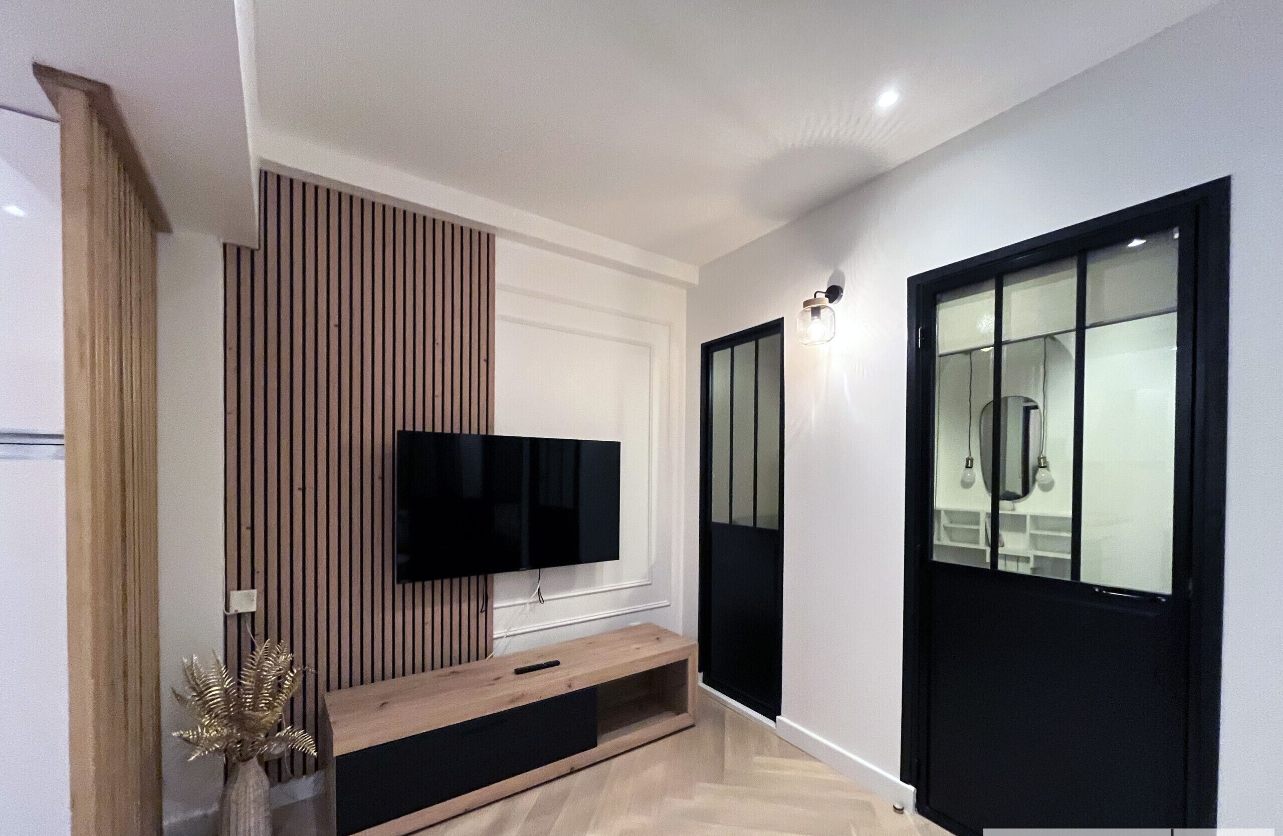 Un pasillo moderno con un televisor de pantalla plana montado en un panel de madera, una puerta de cristal con marco negro y una lámpara en la pared.