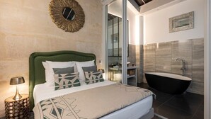 A bedroom with a green headboard, a large bed with patterned bedding, a side table with a lamp, a round mirror on the wall, and a bathroom with a freestanding bathtub. - L'Atelier des Arts in Paris (Paris)