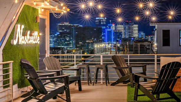 Une terrasse sur le toit, avec des fauteuils Adirondack, un panneau lumineux néon indiquant « Nashville » et une vue panoramique sur la ville.