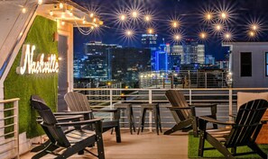 Une terrasse sur le toit, avec des fauteuils Adirondack, un panneau lumineux néon indiquant « Nashville » et une vue panoramique sur la ville.
