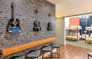 Ein Zimmer mit Ziegelwand, einer hölzernen Bank und drei Gitarren. Es gibt ein Bett mit weißem Kopfteil sowie einen kleinen Tisch mit einer Lampe.