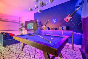 Ein Billardtisch mit Neonreklame, eine Gitarre und eine weitere Neonreklame in einem Raum mit gemustertem Boden und einer Sternenhimmel-Wand.