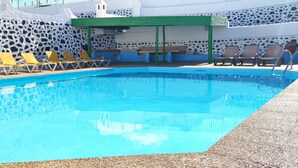 Una piscina con sillas de descanso amarillas y marrones, un muro de piedra y una pérgola verde.