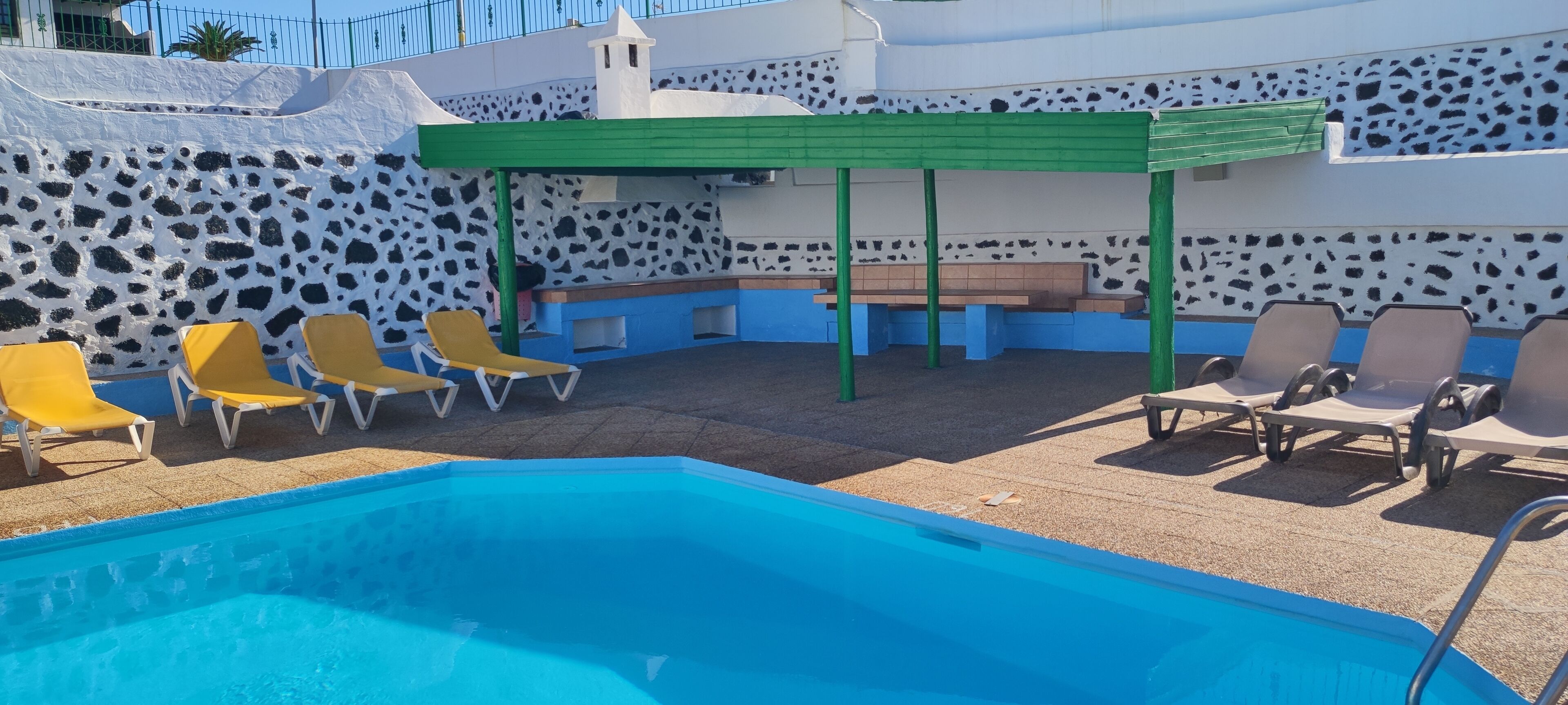 A swimming pool with yellow and brown lounge chairs, a green pergola, and a stone wall with a cow print pattern.