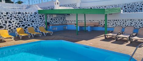 A swimming pool with yellow and brown lounge chairs, a green pergola, and a stone wall with a cow print pattern.
