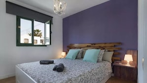 Un dormitorio con una cama, una ventana, una araña y una mesita de noche con una lámpara.