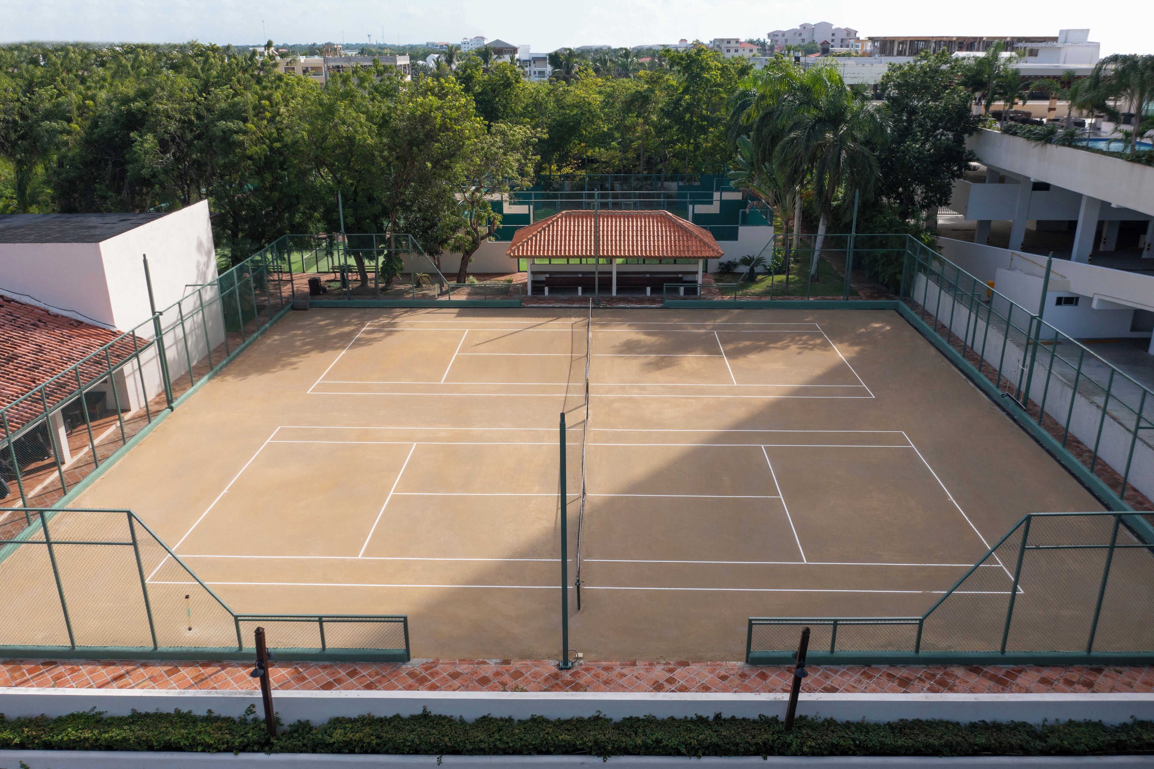 Un court de tennis entouré d’une clôture verte, bordé d’arbres et de bâtiments.