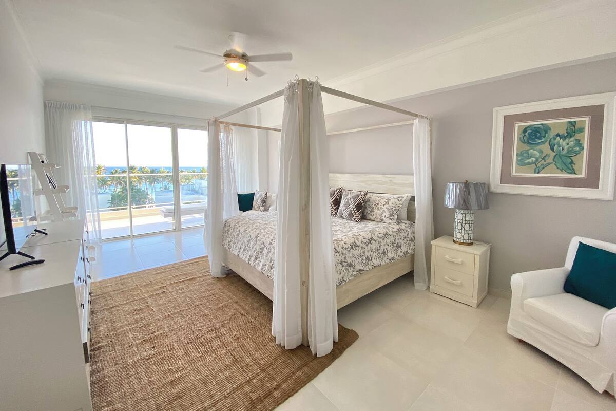 A bedroom with a four-poster bed, a ceiling fan, a bedside table with a lamp, a white chair, and a view of the ocean through large windows.