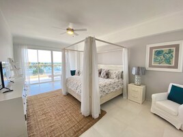 Un dormitorio con cama con dosel, ventilador de techo, mesita de noche con lámpara, una silla blanca y vistas al océano a través de amplias ventanas.
