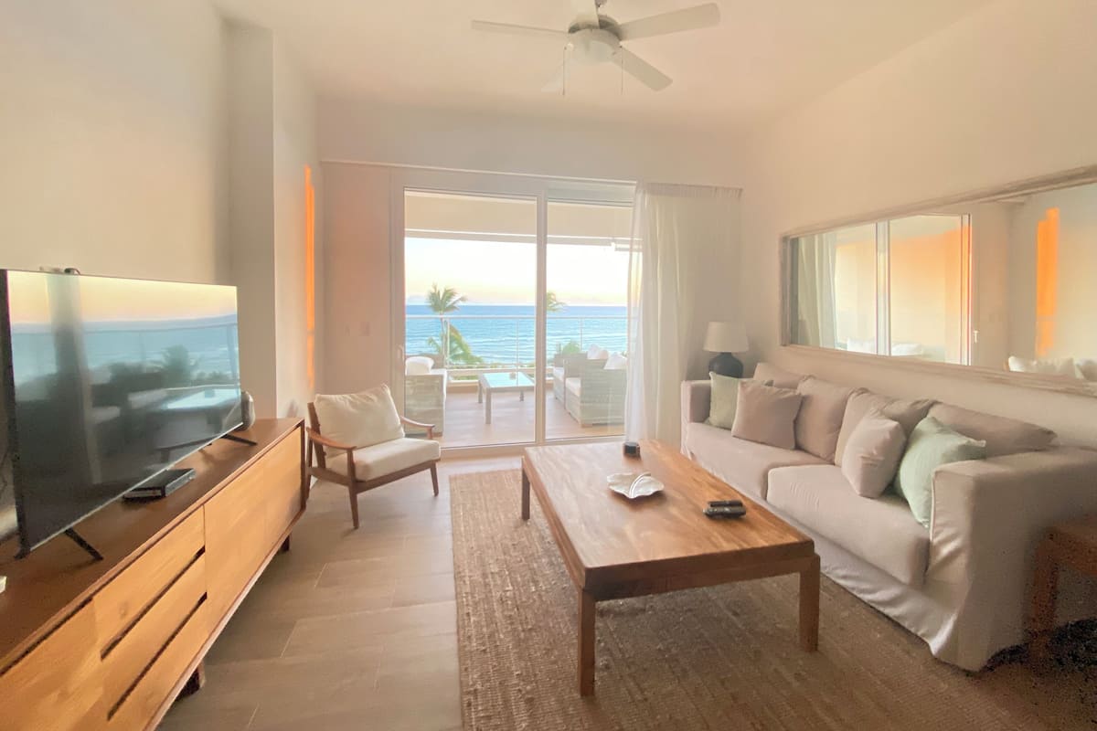 A living room with a television, a wooden coffee table, a sofa, and a chair. There is a large window with a view of the ocean.
