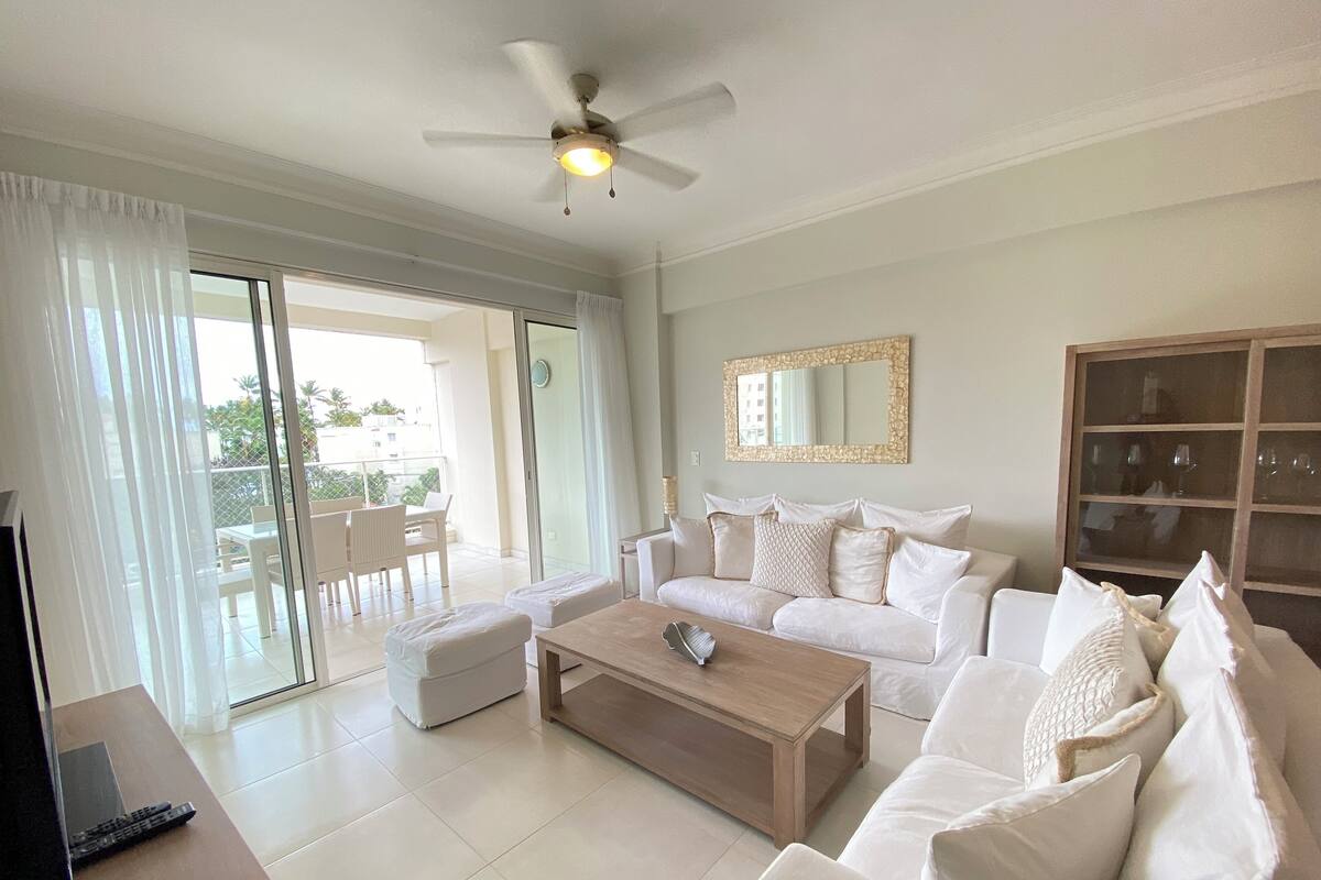 A spacious living room with a white sofa set, a wooden coffee table, a ceiling fan, and a large window with curtains.