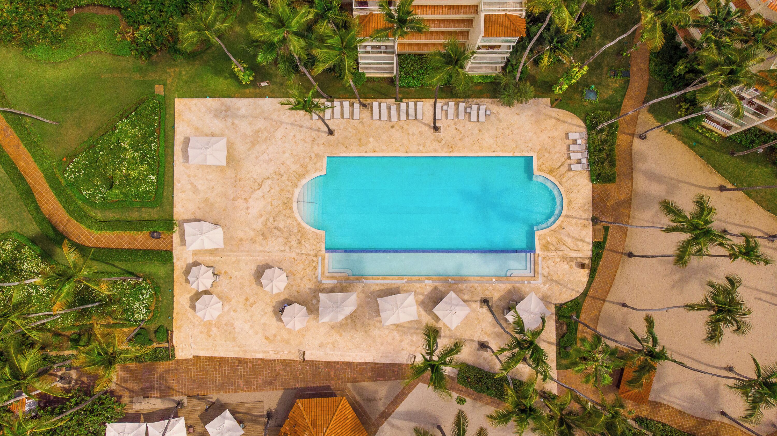 An aerial view of a resort with a central swimming pool, surrounded by palm trees and lounge chairs.