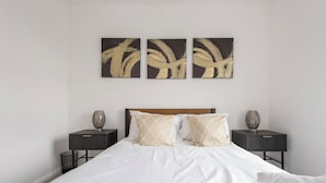 A neatly made bed with white linens and two decorative pillows, flanked by two bedside tables with lamps, in a minimalist room with a plain wall and a single artwork above the bed. - Modern Luxury Appt by Bow Road Station (London)