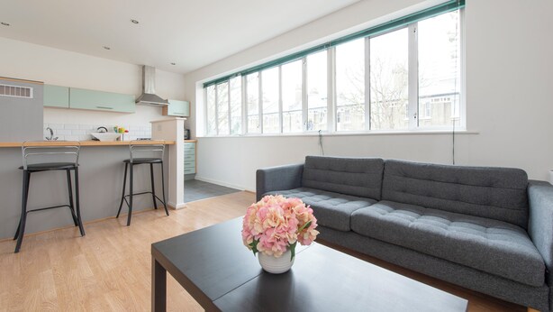 A modern living room with a grey sofa, a coffee table, and a vase of flowers. There are two bar stools and a kitchen area with a window. - Modern Luxury Appt by Bow Road Station (London)