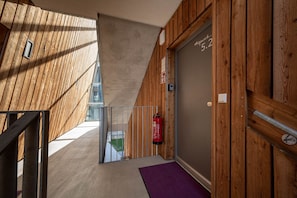 Eine Holzausfachung, ein an der Wand befestigter Feuerlöscher, eine Tür mit einer Nummer und ein violett gefärbter Teppich, der zu einem Balkon führt.