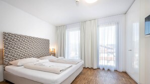 Quarto de hotel com cama, cabeceira com padrão decorativo, piso de madeira e uma janela grande com cortinas finas.