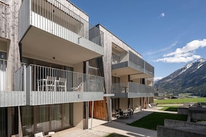 Ein modernes, mehrstöckiges Gebäude mit Balkonen, einer Terrassenfläche mit Gartenmöbeln und einer Bergkulisse im Hintergrund.