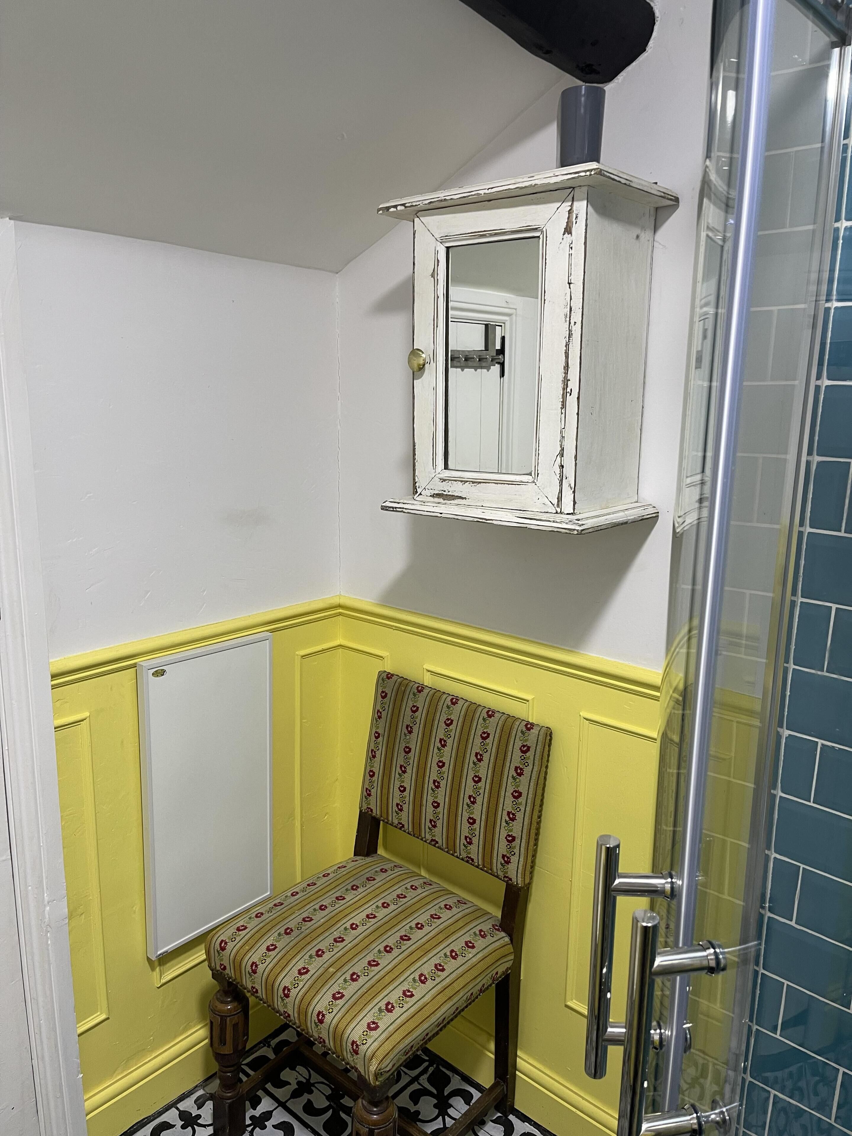 Un baño con paneles inferiores amarillos, una silla de madera tapizada con motivos florales, una puerta blanca y una zona de ducha con azulejos azules.