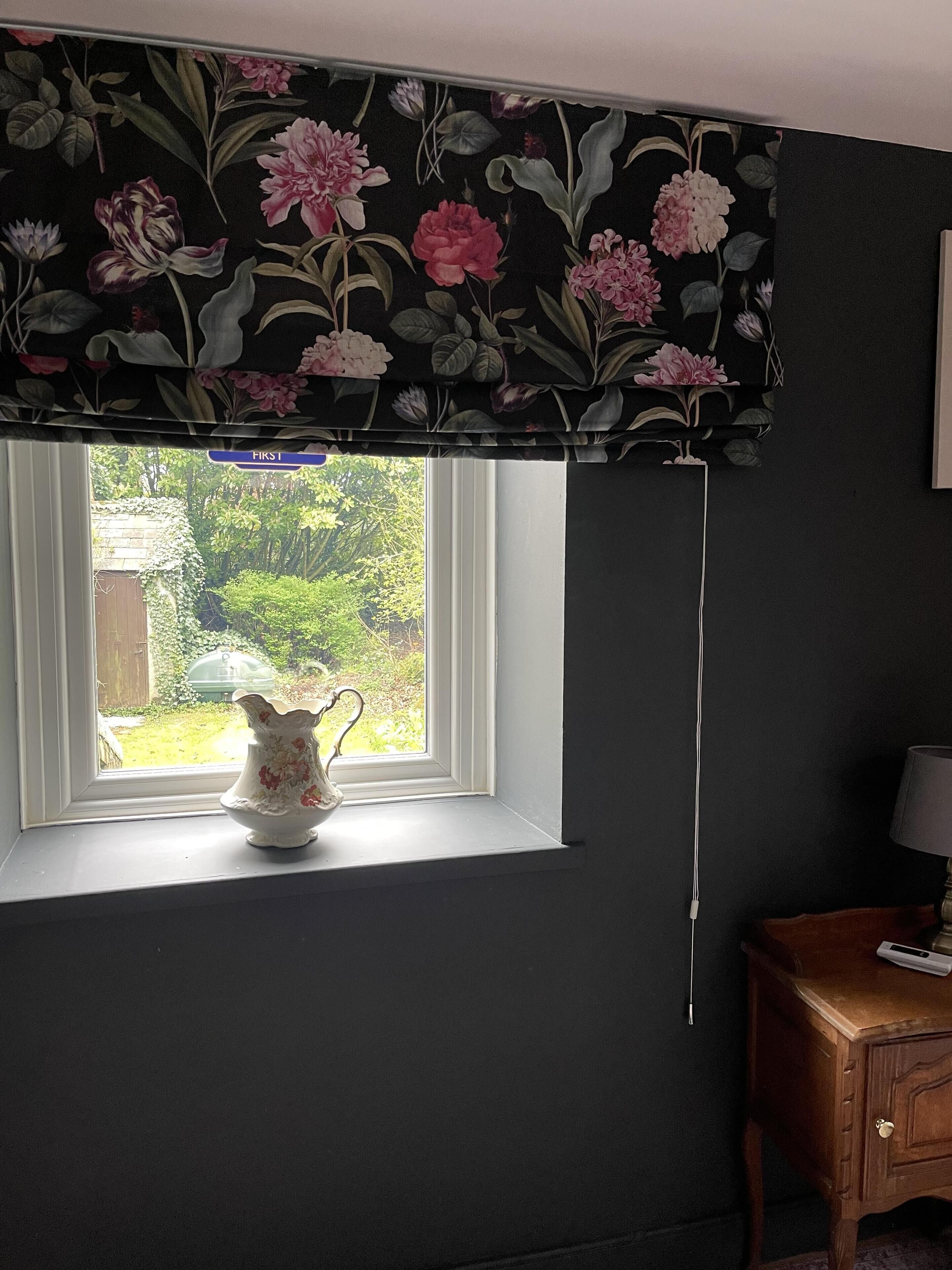 Une pièce avec une fenêtre, une lambrequin à motifs floraux, un vase blanc orné d’un motif de cygne et une table de chevet en bois avec une lampe.