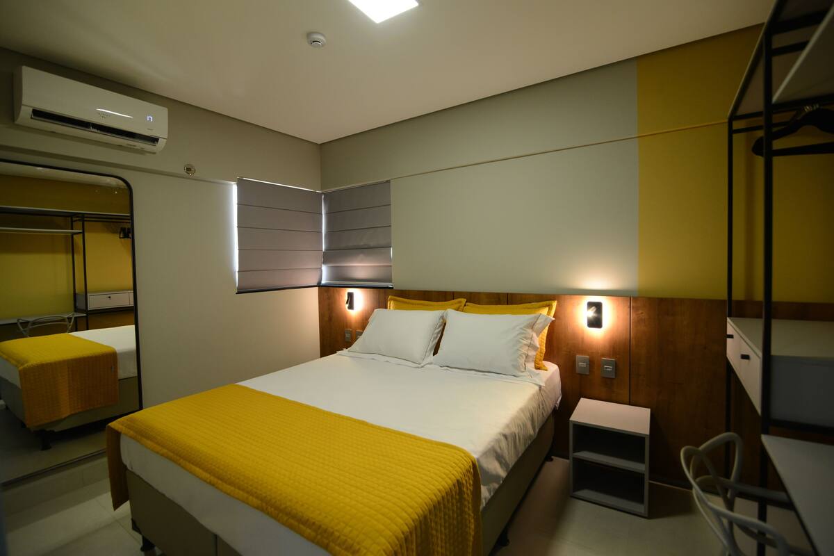 A hotel room with a bed, bedside tables, a mirror, and an air conditioning unit.