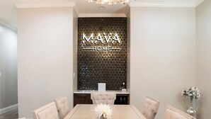 Una zona de menjador amb una taula llarga, sis cadires, un penjó i una paret decorativa amb la inscripció "MAVA HOME".