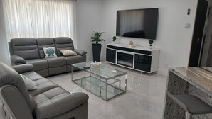A modern living room with a grey sofa, a glass coffee table, a white entertainment unit, and a flat-screen TV mounted on the wall. - CAPE CORAL FLORIDA DREAM $155  last minute  (Cape Coral)