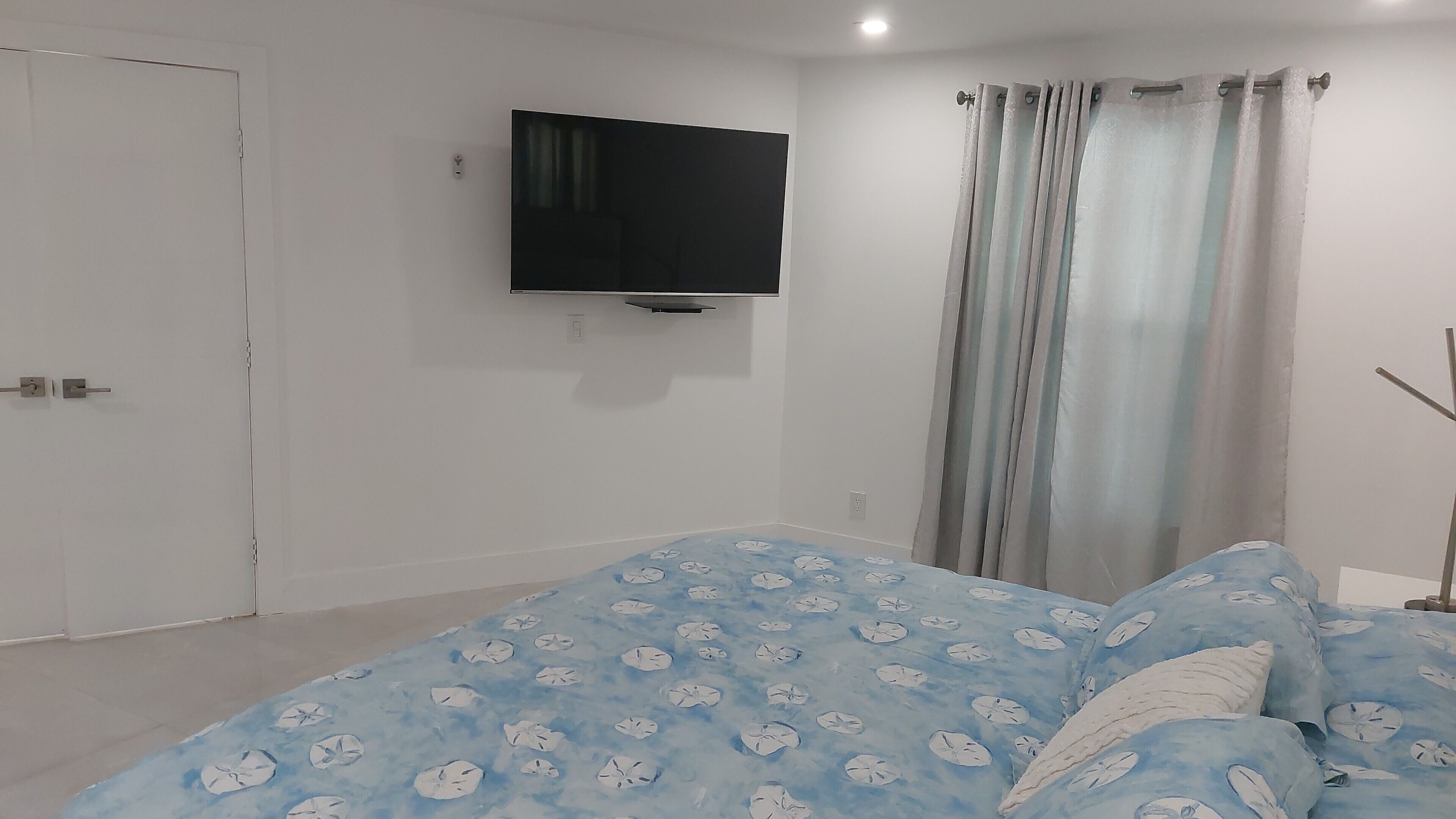 Ein Schlafzimmer mit Bett, einem an der Wand montierten Fernseher, einer Tür und einem Fenster mit Vorhängen.