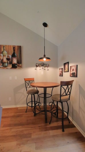 A dining area with a round table and four chairs, a pendant light above, and a painting of wine bottles on the wall.