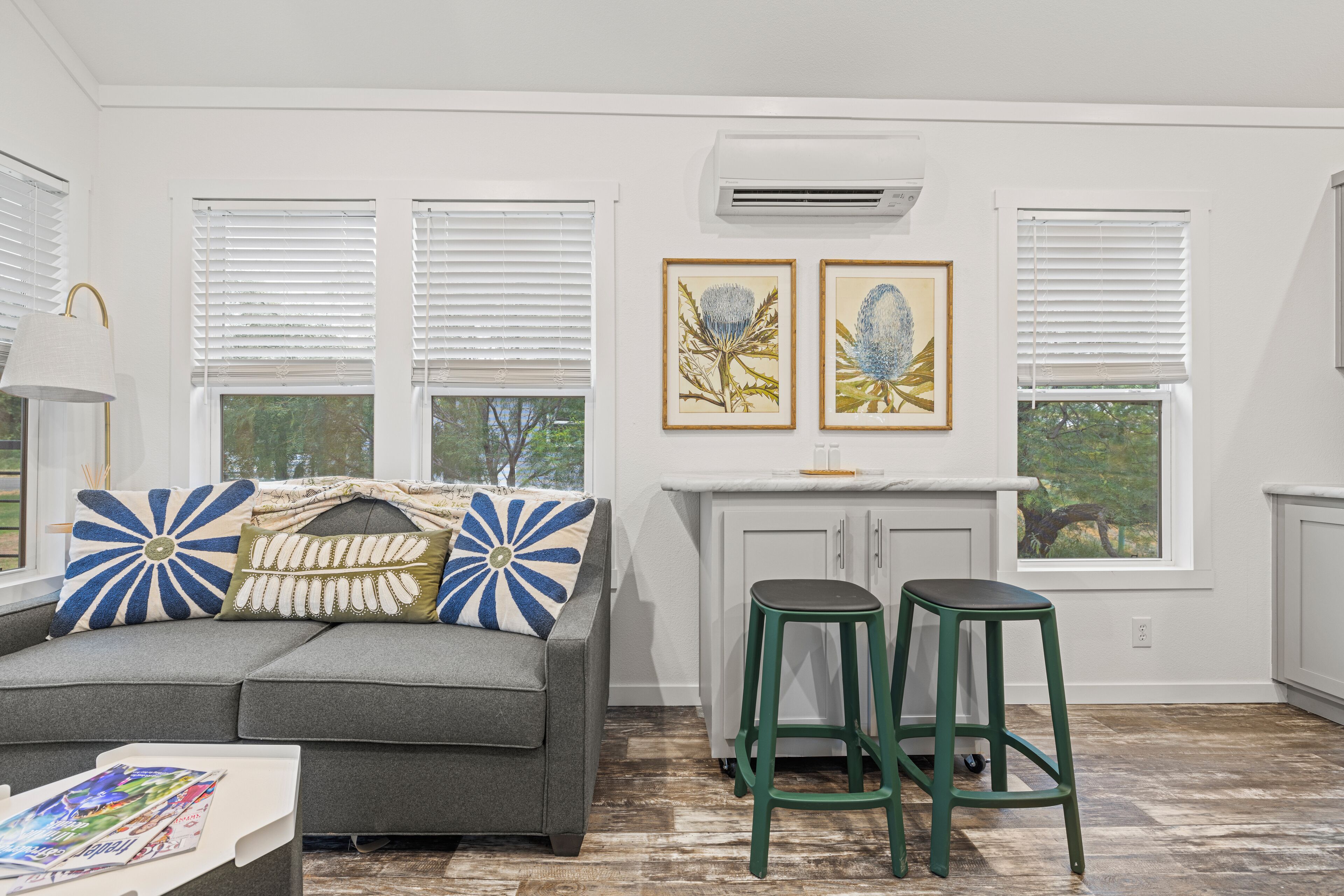Una sala moderna con un sofá gris, dos taburetes verdes y una cocina blanca con ventanal.
