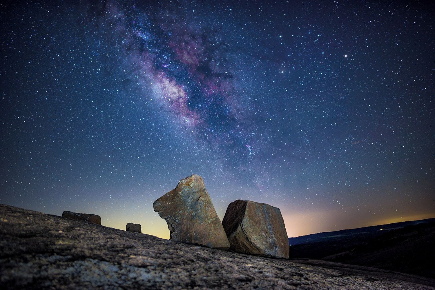 A night sky with stars and the Milky Way, featuring two large rocks in the foreground.