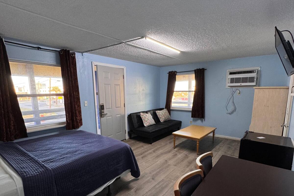 Una habitación con cama, sofá, mesa y escritorio. Se observa una puerta y un equipo de aire acondicionado en la pared.