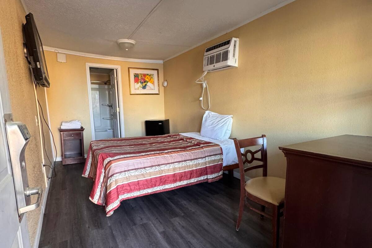 Habitación de hotel con cama, escritorio, silla, unidad de aire acondicionado, televisor y puerta que da a un baño.