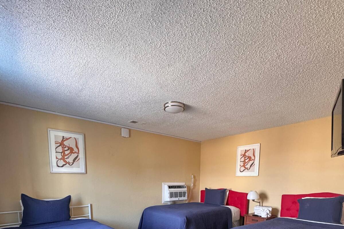 Habitación de hotel con tres camas, ventilador de techo, aire acondicionado y obras de arte en las paredes.