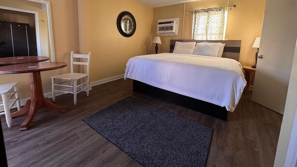 A bedroom with a bed, a desk, a chair, a clock, an air conditioning unit, and a door. - CMI Hotel 1935 Myrtle Beach SC (Myrtle Beach)
